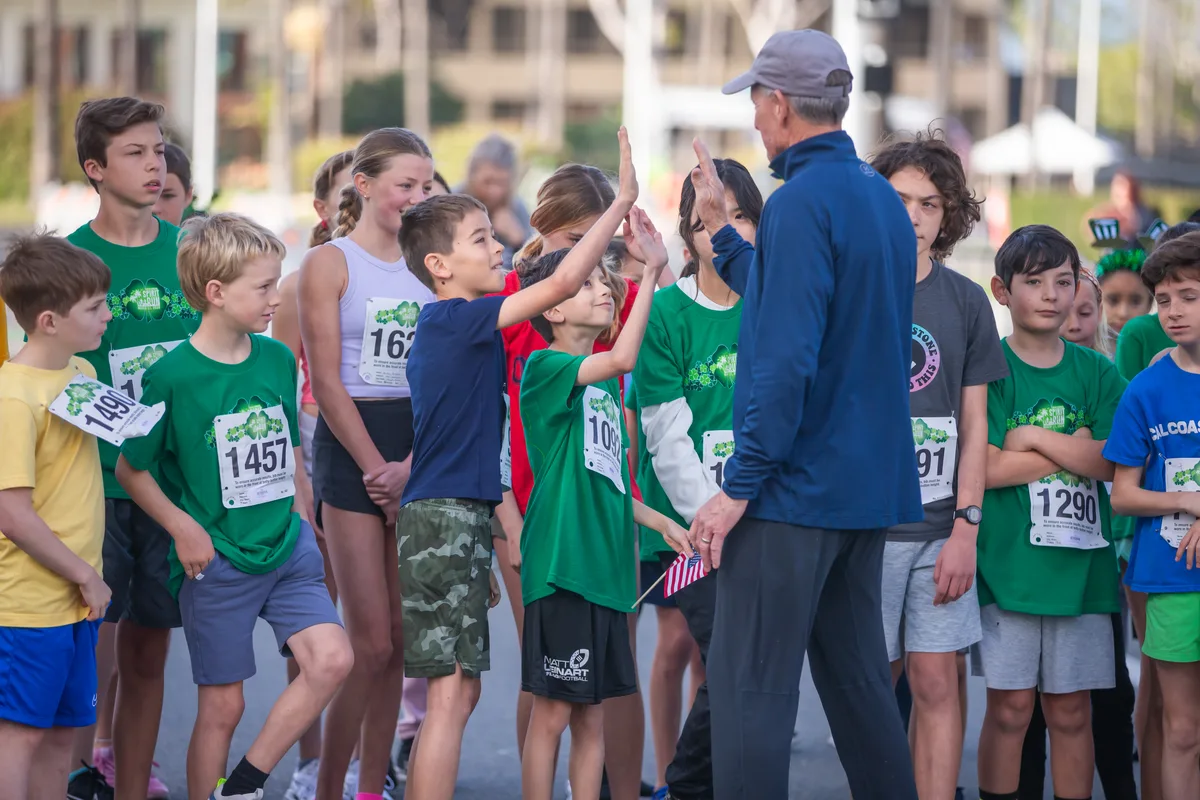 Young runners in Newport Spirit Run shirts exchanging high-fives with a coach at the start line